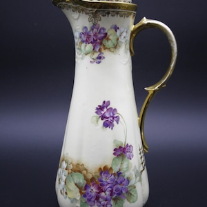 May include: An antique white porcelain pitcher with a gold handle and trim. The pitcher is decorated with hand-painted purple violets and green leaves. The lid is also white with gold accents. A decorative piece for the home.
