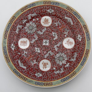 May include: A decorative ceramic plate with a red background and white floral patterns. The plate has a patterned border with blue, yellow, and white accents. Four white circles with Chinese characters are evenly spaced.