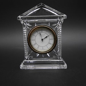 May include: A clear crystal clock with a gold-tone bezel and a white clock face. The clock is encased in a clear crystal structure with a peaked roof and decorative detailing. The clock face reads "Waterford Crystal".