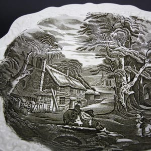 May include: A white, scalloped-edge plate with a black and white transferware scene. The scene depicts a rural landscape with a cottage, trees, figures, and a church in the distance. The plate has a detailed, vintage aesthetic.