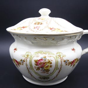 May include: A white porcelain chamber pot with a lid and handle. The pot is decorated with floral patterns in pink, yellow, and green, and has gold trim. The lid has a small knob on top.