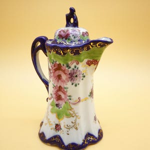 Peut inclure: Une cafetière en porcelaine blanche avec un motif floral. Le pot présente des fleurs roses, violettes et vertes, avec des accents dorés et un bord bleu. La poignée et le couvercle sont également bleus.