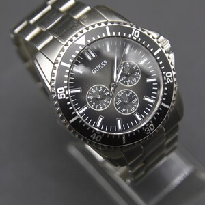 May include: A silver Guess watch with a black bezel and a dark grey face. The watch has three sub-dials and a date display. The watch strap is made of silver metal links.