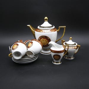 May include: A white porcelain tea set with gold accents, featuring a teapot, creamer, sugar bowl, cups, and saucers. The set has a decorative design with a red and gold pattern and a Medusa head emblem.
