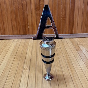 May include: A silver-colored bottle stopper with a large letter "A" on top. The stopper has a conical shape with two black rubber rings and a flared base. The text "Hand wash only Made in China" is printed on the stopper.