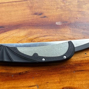 May include: A folding pocket knife with a black handle and a silver blade. The handle has a textured grip and is accented with silver rivets. The blade is curved and has a sharp edge. The knife is lying on a wooden surface.