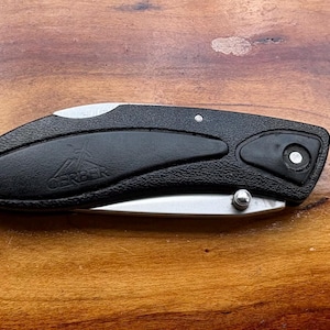 May include: A black folding knife with a silver blade and a textured handle. The knife is closed, with the blade tucked inside the handle. The handle has a curved design and a small silver button. The knife is on a wooden surface.