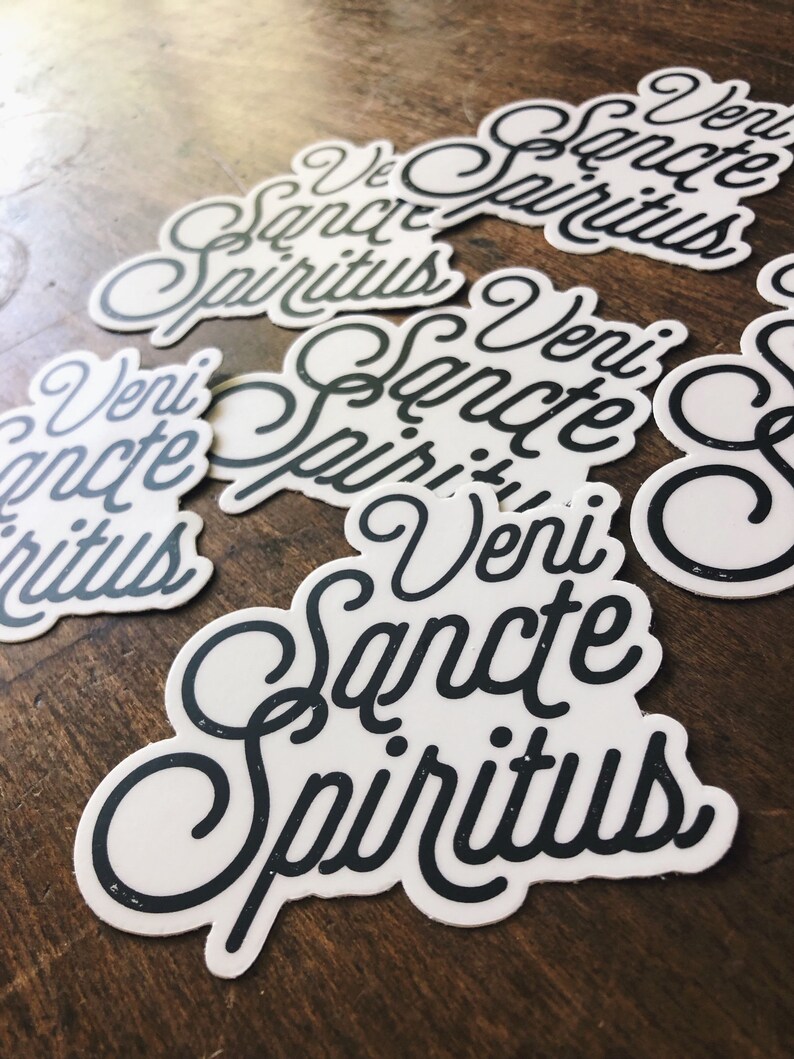 Puede incluir: Un conjunto de cuatro pegatinas blancas con letras negras que dicen "veni sancte spiritus". Las pegatinas est&aacute;n sobre una superficie de madera.