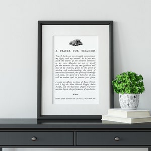 May include: Framed print with the text "A Prayer for Teachers" in black font. The print is displayed on a dark gray table, with a small potted plant and books. The frame is black.