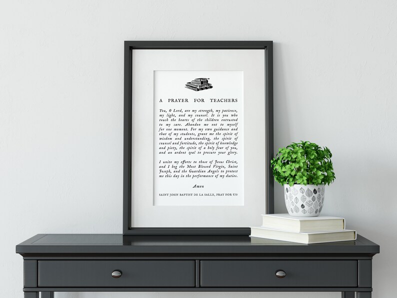 May include: Framed print with the text "A Prayer for Teachers" in black font. The print is displayed on a dark gray table, with a small potted plant and books. The frame is black.