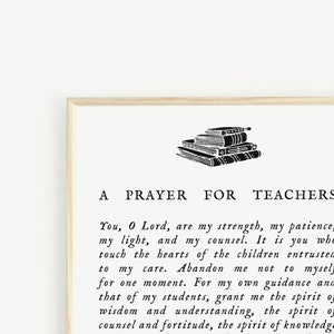 May include: A framed print with the text "A Prayer for Teachers" in black font. The print features a stack of books at the top. The background is a light cream color, and the frame is a light wood color.