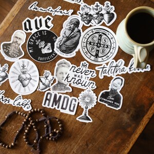 Puede incluir: Una colecci&oacute;n de pegatinas en blanco y negro con citas e im&aacute;genes religiosas e inspiradoras. Las pegatinas est&aacute;n sobre una superficie de madera con una taza de caf&eacute; y un rosario.