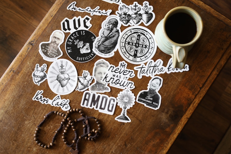 Puede incluir: Una colecci&oacute;n de pegatinas en blanco y negro con citas e im&aacute;genes religiosas e inspiradoras. Las pegatinas est&aacute;n sobre una superficie de madera con una taza de caf&eacute; y un rosario.