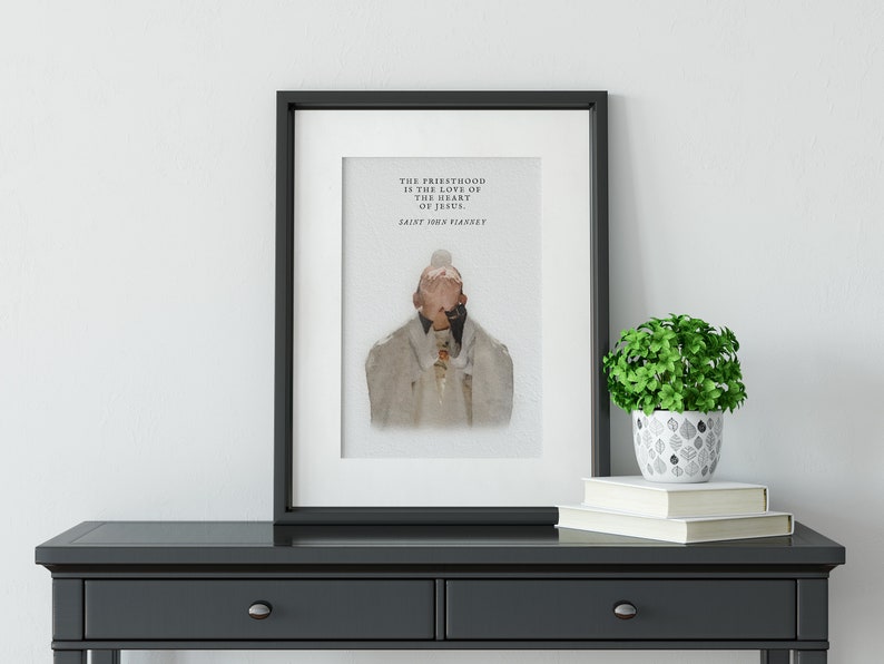 Op de afbeelding: Een zwart ingelijst afdruk met een aquarel-illustratie van een persoon in een witte gewaden met hun handen voor hun gezicht. De tekst op de afdruk luidt "Het priesterschap is de liefde van het hart van Jezus. Heilige Johannes Vianney."