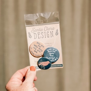 May include: Three round pins with inspirational quotes and a bird design. The pins are blue and pink with white lettering. The text on the pins reads "You are not who they say you are. St. John Paul II", "You are worth more than many sparrows", and "Totally Himself".