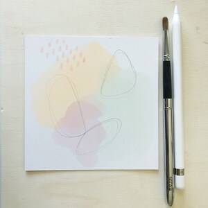 Op de afbeelding: Een witte kaart met abstracte aquarelvormen in tinten roze, geel en groen. De kaart heeft dunne zwarte lijnen erop getekend. Twee zilveren gereedschappen, een stylus en een penseel, liggen naast de kaart.