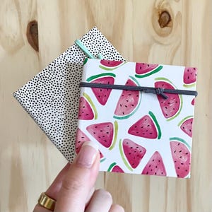Può includere: Due piccoli quaderni avvolti in carta decorativa. Un quaderno ha un motivo a pois neri e bianchi. L'altro quaderno ha uno sfondo bianco con un motivo ripetuto di angurie rosse e verdi.