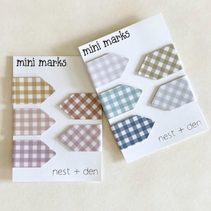 May include: Two white display cards labelled mini marks and nest + den, each holding three arrow-shaped page markers with a gingham pattern. The markers feature various muted colours including mustard, pink, lavender, sage green, and blue shades.