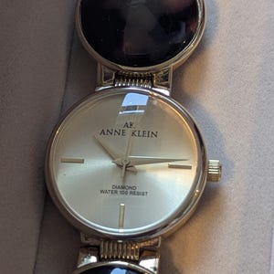 Può includere: Orologio Anne Klein color oro con quadrante rotondo e cinturino dorato. Il quadrante dell'orologio presenta il logo Anne Klein e le parole "Diamond Water 100 Resist". Il cinturino presenta maglie circolari nere e dorate.