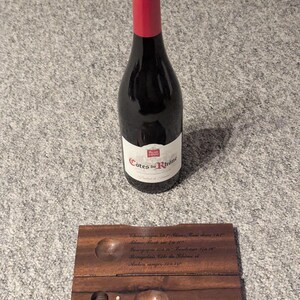 Könnte beinhalten: Eine dunkle Weinflasche mit rotem Verschluss und weißem Etikett mit der Aufschrift "Cotes du Rhone" steht über einem Weinthermometer aus Holz und einem kleinen Holzhalter.