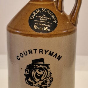 Könnte beinhalten: Brauner Steinzeugkrug mit Henkel, mit der Aufschrift "Farm Scrumpy" und "Countryman Cider". Der Krug hat ein Fassungsvermögen von 55cl. Der Krug hat einen dunkelbraunen Deckel und einen cremefarbenen Körper.