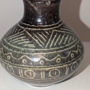 Könnte beinhalten: Kleine, runde Keramikvase mit dunkelbraunem Oberteil und grauem Körper. Der Körper ist mit weißen geometrischen Mustern verziert, darunter Linien, Kreise und augenähnliche Formen. Die Vase hat eine glatte, glänzende Oberfläche.