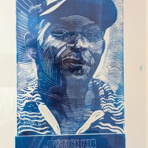 May include: Blue and white linocut print portrait of a person wearing a cap and patterned shirt. The text "JUSTICE FOR CHRISTOPHER ALDER" is at the bottom. The artwork is framed in white.