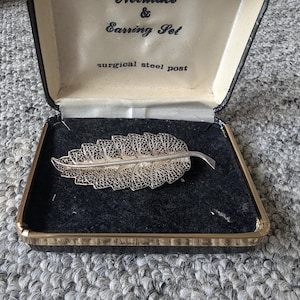 Op de afbeelding: Een zilverkleurige filigraan bladbroche in een zwart fluwelen sieradendoosje. De doos is open en toont het ingewikkelde ontwerp van de broche. Op het deksel staat "Necklace & Earring Set" en "surgical steel post".