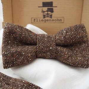 Könnte beinhalten: Eine braune Tweed-Schleife und ein passender Stoffstreifen, die auf weißem Stoff liegen. Die Schleife mit ihrem strukturierten Muster ist das zentrale Element. Im Hintergrund ist eine Pappschachtel mit dem Logo "fliegenshon" zu sehen.