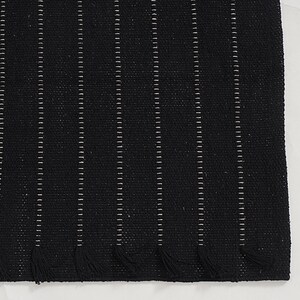 May include: A black woven rug with vertical white stripes and fringe at the bottom. The rug has a textured appearance and is likely made of natural fibers. The design is simple and modern, suitable for various interior styles.