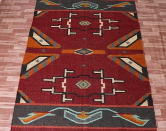 May include: A rectangular, red area rug with a geometric pattern. The rug features a central design with white, black, orange, and gray shapes. The border is dark gray with orange and teal accents. The rug has a fringed edge.
