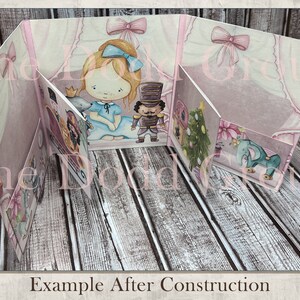 May include: A pink and white papercraft pop-up card with a girl, a nutcracker, a Christmas tree, and an elephant. The card is open and the pop-up elements are visible. The card is on a wooden surface.  Example After Construction