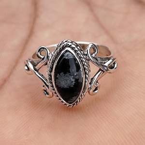Peut inclure: Une bague en argent ornée d'une pierre noire de forme marquise avec des inclusions blanches. Le sertissage présente un bord en corde torsadée autour de la pierre et des volutes décoratives sur les côtés de l'anneau, créant un design détaillé.