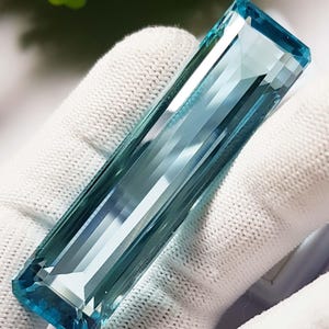 Może przedstawiać: Prostokątny, fasetowany kamień szlachetny akwamaryn o jasnoniebieskim odcieniu. Kamień trzymany jest w białej rękawicy, co podkreśla jego przejrzystość i szlif. Fasetki odbijają światło, tworząc efekt iskrzenia. Idealny do tworzenia biżuterii.