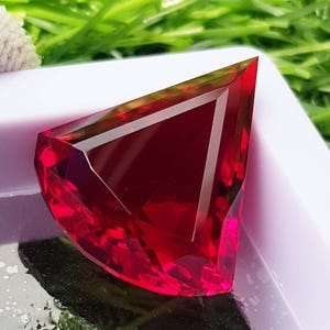 May include: A faceted, triangular-shaped gemstone with a deep red hue. The gem has a polished surface with multiple facets that reflect light, creating a brilliant sparkle. The gem is set against a white background with green foliage.