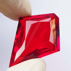May include: A faceted, kite-shaped gemstone in a vibrant red hue. The gem is held by a gloved hand, showcasing its sharp angles and internal reflections. The gemstone's cut allows light to refract, creating a brilliant display.