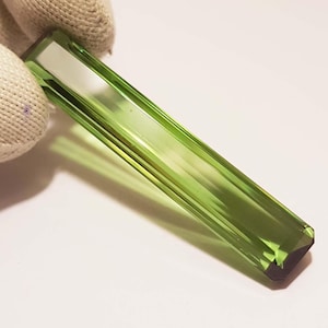 May include: A rectangular, faceted, light green gemstone. The gem is held by a gloved hand, highlighting its polished surfaces and internal clarity. The gemstone's colour is a vibrant, translucent green.