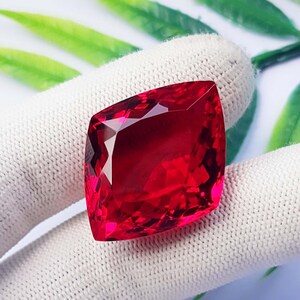 Può includere: Una gemma rosso intenso e vibrante con taglio a cuscino e superficie sfaccettata. La pietra mostra elevata chiarezza e brillantezza, riflettendo la luce attraverso le sue sfaccettature geometriche su uno sfondo di tessuto bianco testurizzato.