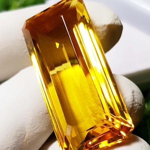 Könnte beinhalten: Ein rechteckiger, facettierter Edelstein mit leuchtend gelber Farbe. Der Edelstein hat eine polierte Oberfläche und ein klares, transparentes Aussehen. Der Edelstein ist wahrscheinlich ein Citrin oder eine ähnliche Kristallart. Der Edelstein wird auf einer weißen Oberfläche präsentiert.