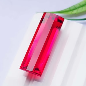 Puede incluir: Una piedra preciosa rectangular facetada de un color rojo intenso y vibrante. La piedra presenta un corte tipo baguette largo y delgado con bordes afilados y limpios, y una superficie altamente pulida y reflectante que capta la luz.