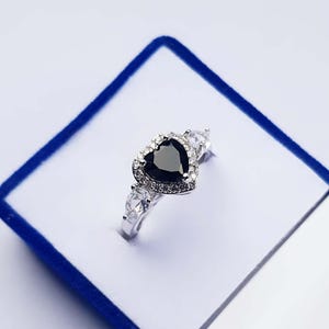 May include: A silver-toned ring featuring a central heart-shaped black gemstone surrounded by a halo of small clear stones. The band includes a pear-shaped clear stone on each side, set in a metallic frame with a polished finish.