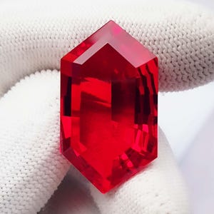 May include: A vibrant, transparent red gemstone cut into a hexagonal shape with multiple facets. The stone displays a deep crimson hue and sharp, geometric edges, reflecting light across its polished surface to create a clear, crystalline appearance.