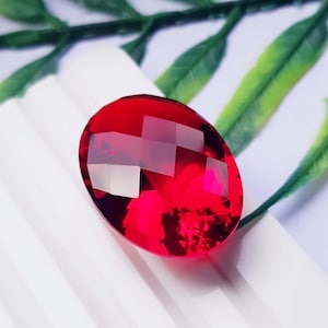Könnte beinhalten: Ein leuchtend roter, oval geformter Edelstein mit einer facettierten Oberfläche, die das Licht reflektiert. Der Stein zeigt einen tiefen, gesättigten Karmesinton mit geometrischen Schliffen auf der Oberseite, was für ein brillantes Funkeln sorgt.