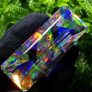 May include: A multi-coloured, rectangular gemstone, displaying a spectrum of hues such as blue, green, yellow, and orange. The stone is held in a black glove, with green foliage in the background. The cut of the stone creates many facets, reflecting light.
