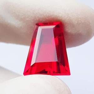 May include: A faceted, trapezoid-shaped, vibrant red gemstone. The gem is held against a white background, showcasing its sharp angles and internal clarity. The intense color and precise cuts highlight its potential for jewelry design.