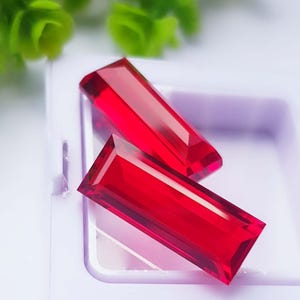 May include: Two rectangular, faceted, vibrant red gemstones are positioned inside a white plastic display case. The stones feature sharp, clean edges and a polished surface that reflects light, showcasing a deep, saturated crimson color throughout.