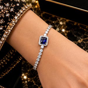 Può includere: Un bracciale tennis color argento con una pietra blu rettangolare centrale circondata da un alone di piccole pietre chiare, con una fila continua di pietre rotonde chiare lungo la fascia. La scatola mostra il testo LUXURY TIMELESS ELEGANCE.