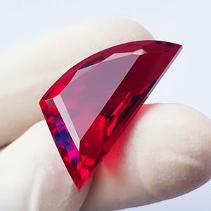 Op de afbeelding: Een gefacetteerde, levendige rode edelsteen met een unieke, hoekige vorm. De gepolijste oppervlakken van de edelsteen reflecteren licht, wat de helderheid en rijke kleur benadrukt. De edelsteen wordt tegen een witte achtergrond gehouden.