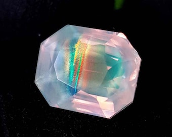 May include: A faceted gemstone with a rectangular shape. The gem has a pale pink hue with internal rainbow-colored iridescence. The gem is set against a black background, highlighting its clarity and brilliance.