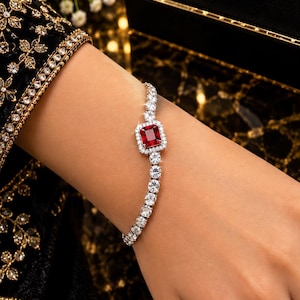 Puede incluir: Una pulsera de tenis en tono plateado con una piedra preciosa roja rectangular central rodeada por un halo de pequeñas piedras claras, con una línea continua de piedras redondas claras a lo largo de la banda, presentada en una caja negra con LUXURY.